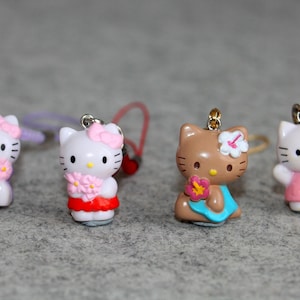 May include: Four Hello Kitty charms in various outfits. Two white figures have pink bows and hold flowers. One brown figure wears a blue dress and holds a flower. The last white figure has an orange bow and a yellow flower.