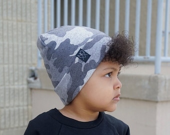 May include: A young person wearing a gray and black camouflage beanie with a black logo. The beanie has a fold-over cuff. The person is wearing a black sweatshirt with the word "be" printed on the front.