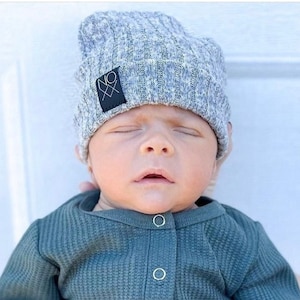 May include: A baby wearing a gray knitted beanie with a black logo and a green waffle knit onesie.