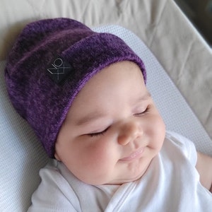 May include: A baby wearing a purple beanie with a black geometric logo. The baby is sleeping and has a white shirt on.