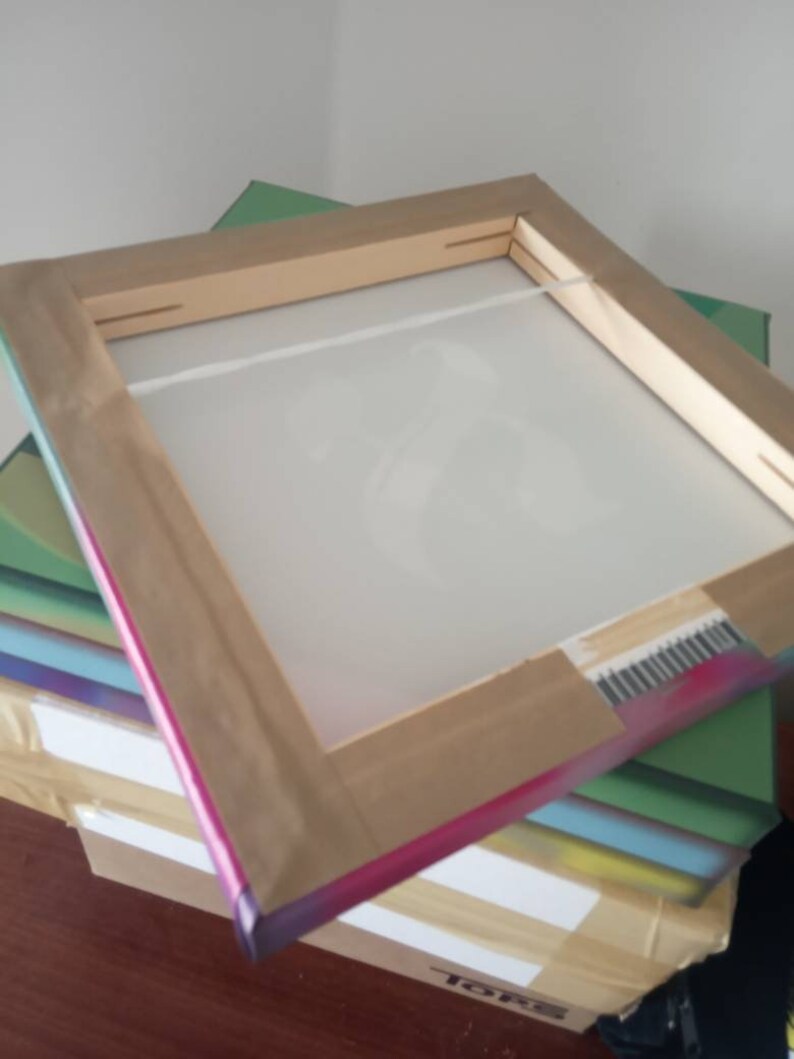 May include: A wooden frame with a white canvas stretched over it. The frame is wrapped in a rainbow-colored tape. The frame is sitting on top of a stack of boxes.