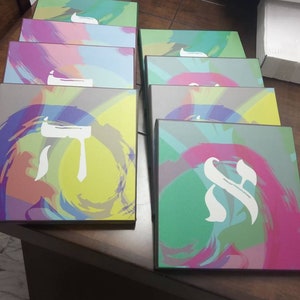 May include: A stack of colorful boxes with Hebrew letters on them. The boxes are decorated with abstract brushstrokes in shades of green, blue, pink, and yellow.