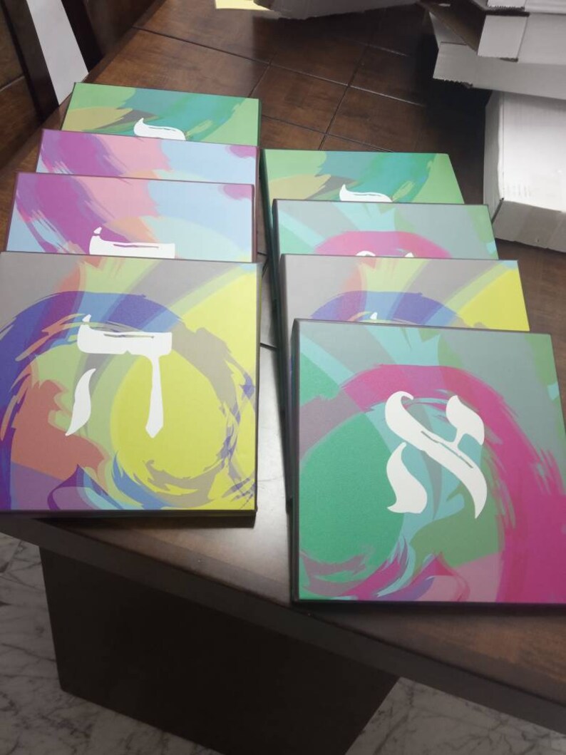 May include: A stack of colorful boxes with Hebrew letters on them. The boxes are decorated with abstract brushstrokes in shades of green, blue, pink, and yellow.