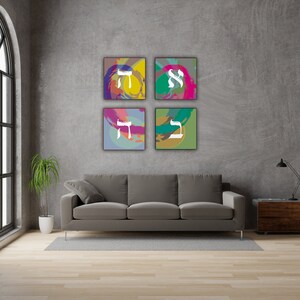 May include: Four square abstract art prints with Hebrew letters in white on a gray background. The prints are arranged in a grid pattern on a gray wall above a gray couch.