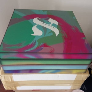 May include: A stack of four canvases with abstract paintings in shades of green, blue, pink, and purple. The top canvas features a white stylized letter "S" in the center.