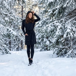 May include: A black hooded long-sleeve dress with a drawstring waist and two front pockets, paired with black leggings and black ankle boots. The garment features a fitted silhouette and a hood, providing full coverage in a cold outdoor environment.