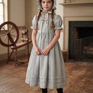 May include: A light gray dress with short puff sleeves, a gathered skirt, and a matching bonnet with a tie closure. The dress features a square neckline with a small bow detail and horizontal pleats along the hemline, paired with black boots.