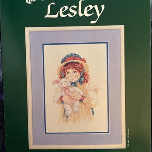 Könnte beinhalten: Kreuzstich-Anleitungsheft mit dem Titel "Lesley" von Jan Hagara. Das Bild zeigt eine gerahmte Kreuzsticharbeit eines Mädchens mit einem Stoffhasen. Das Heft ist grün mit Text, einschließlich Preis und Herausgeber, Johnson Creative Arts.