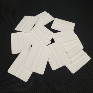 May include: A pile of white plastic squeegees with a textured surface. The squeegees are used for applying vinyl decals and other adhesive materials.