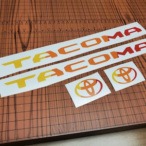 May include: Two white vinyl decals with the word "TACOMA" in yellow and orange letters. Two smaller white vinyl decals with a yellow and orange Toyota logo.