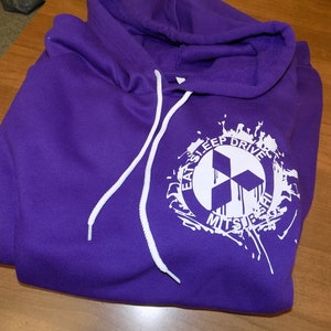 Może przedstawiać: Fioletowa bluza z kapturem z białym nadrukiem graficznym w kształcie koła z tekstem "Eat Sleep Drive Mitsubishi" i stylizowanym logo Mitsubishi.