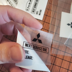 Może przedstawiać: Przezroczysta naklejka z tworzywa sztucznego z czarnym logo Mitsubishi i tekstem "NEXT SERVICE DUE". Naklejka ma białe tło i czarny tekst dla "DATE:" i "MILEAGE:".