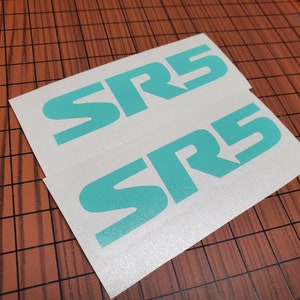 Puede incluir: Dos pegatinas blancas con el texto "SR5" en letras de color turquesa. Las pegatinas están sobre una superficie marrón.