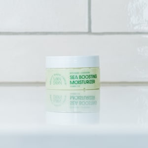 May include: A white jar with a green label that reads "MADE SIMPLE MIN SKIN" and "SEA BOOSTING MOISTURIZER" with algae and cloudberry. The jar is sitting on a white surface.