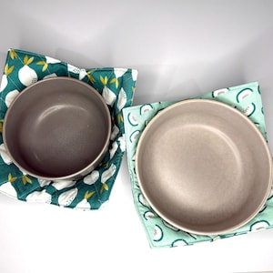 May include: Two ceramic bowls rest on quilted bowl cozies. One bowl is gray, the other is beige. The cozies feature a white and yellow lemon pattern on a teal background, and a white and green melon pattern on a light green background.