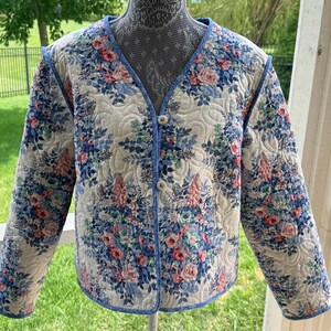 May include: A quilted jacket featuring a cream-colored base with a floral pattern in shades of blue, pink, and green. The garment has a V-neckline, long sleeves, and two round buttons down the front, all finished with a blue patterned fabric trim.