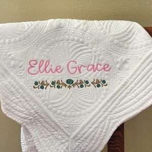 K&ouml;nnte beinhalten: Wei&szlig; gesteppte Babydecke mit gesticktem Text "Ellie Grace" in Rosa. Die Decke hat ein detailliertes Blumendesign und eine dekorative Bord&uuml;re in Braun und Blaugr&uuml;n. Die Decke ist &uuml;ber einen Holzstuhl drapiert.