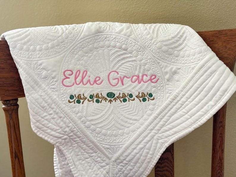 K&ouml;nnte beinhalten: Wei&szlig; gesteppte Babydecke mit gesticktem Text "Ellie Grace" in Rosa. Die Decke hat ein detailliertes Blumendesign und eine dekorative Bord&uuml;re in Braun und Blaugr&uuml;n. Die Decke ist &uuml;ber einen Holzstuhl drapiert.