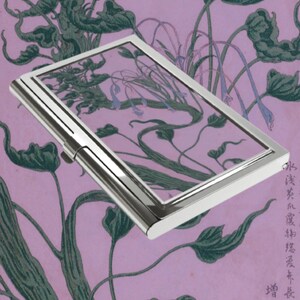 May include: A rectangular silver-toned metal business card case with a polished finish. The case features a hinged lid and a slim profile. The surface of the case reflects a background pattern of dark green botanical illustrations on a light purple field.