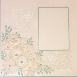 May include: A white scrapbook page with a green border around a blank rectangle in the center. The page is decorated with white lace, white flowers, and green leaves.