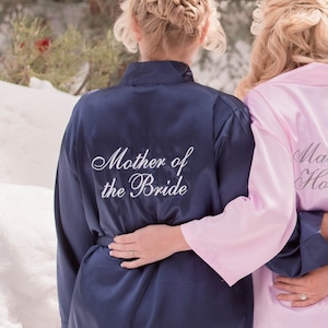 May include: Two women wearing matching satin robes. The woman in the front is wearing a navy blue robe with the text "Mother of the Bride" embroidered on the back. The woman in the back is wearing a pink robe.