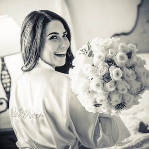 Pu&ograve; includere: Una sposa con una vestaglia di raso bianco con il testo "Bride's Party" ricamato sul davanti. Tiene un bouquet di rose bianche.