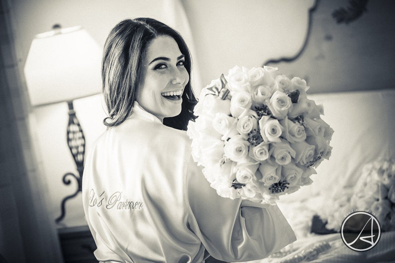 Pu&ograve; includere: Una sposa con una vestaglia di raso bianco con il testo "Bride's Party" ricamato sul davanti. Tiene un bouquet di rose bianche.