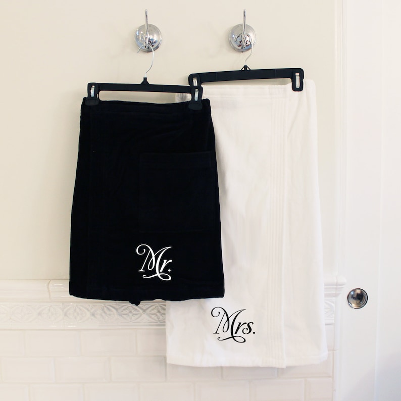 May include: A set of two white bath towels with black embroidery. The towels are hanging on hooks. The towel on the left has "Mr." embroidered on it. The towel on the right has "Mrs." embroidered on it.