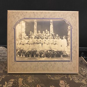 May include: A framed black and white photograph of a group of people, likely nurses, standing in front of a building. They are holding bouquets of flowers and are smiling. The photo is framed in a brown frame with a blue border.