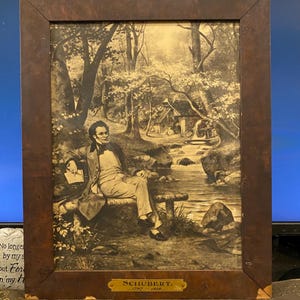 May include: A framed sepia-toned print of a man sitting on a bench in a forest setting. The man is wearing a suit and has a thoughtful expression on his face. The print is titled "SCHUBERT. 1797 - 1828".