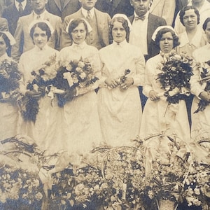 May include: A group of women in white dresses and caps stand in front of a group of men in suits. They are holding bouquets of flowers. The women are smiling and looking at the camera.
