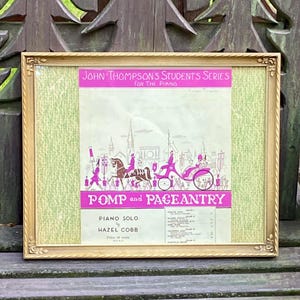 May include: A framed vintage sheet music cover titled John Thompson's Students Series for the Piano, Pomp and Pageantry by Hazel Cobb. The print features a pink and white illustration of a horse-drawn carriage in a city, set in a gold frame.