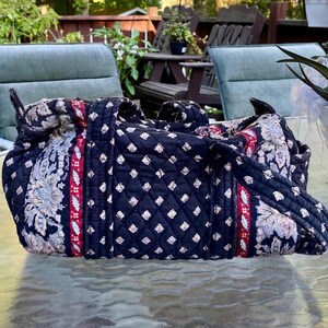 May include: A quilted fabric duffel bag in a dark navy blue color featuring a repeating white floral pattern, accented by vertical bands of red and white decorative trim. The bag has two sturdy fabric handles and a rectangular, structured shape.