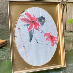 May include: A framed watercolor painting featuring a bird perched on a branch with red flowers. The artwork is set against a white background within an oval shape, framed by a gold-colored frame. The painting has a traditional Asian art style.