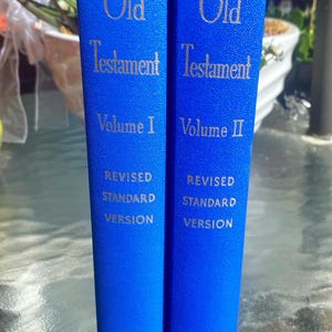 May include: Two bright blue books with gold lettering on the spines. The text reads Old Testament Volume I and Volume II, Revised Standard Version, and Nelson. The books have a textured cover finish and are positioned vertically side by side.
