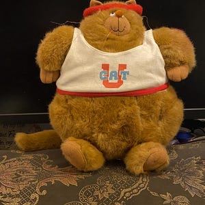 May include: A plush brown cat wearing a white tank top with "CAT" in blue and red lettering. The cat has a red headband and a red band around the waist. The cat is sitting down with its tail extended.