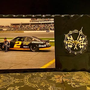 May include: Black framed clock with a racing theme. The left side features a photo of a race car with the number 2 and the words "Miller Genuine Draft." The right side has a clock with checkered flags.