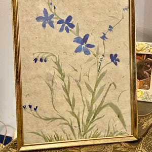 May include: A framed watercolor painting featuring blue flowers and green stems against a beige background. The artwork is enclosed in a gold-colored frame, adding a touch of elegance to the floral design. The painting evokes a sense of nature and tranquility.