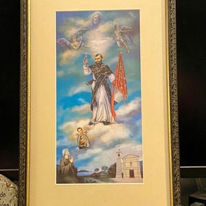 May include: Framed print of a religious image depicting a man in a brown robe holding a cross and a red flag, surrounded by angels and a church in the background.