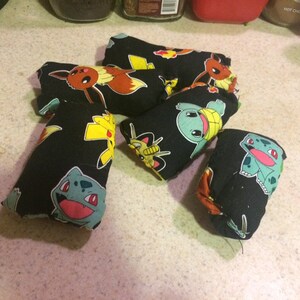 May include: Six black fabric pouches with different Pokemon characters printed on them. The pouches are filled with something soft and are likely used as stress balls or fidget toys.