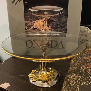 May include: A two-tiered serving compote with a clear glass top and a gold-colored rim. The base is silver with gold rose accents. The text "Golden Damask Rose Compote" is visible on the packaging. The brand name "ONEIDA" is also visible.