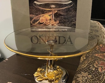 May include: A two-tiered serving compote with a clear glass top and a gold-colored rim. The base is silver with gold rose accents. The text "Golden Damask Rose Compote" is visible on the packaging. The brand name "ONEIDA" is also visible.