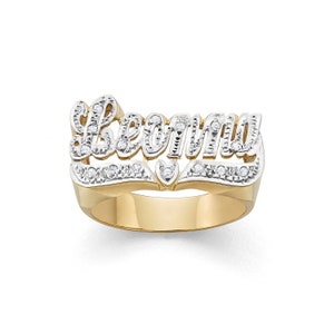 Könnte beinhalten: Ein goldfarbener Ring mit dem Namen "Berny" in einem mehrschichtigen Design. Die obere Schicht zeigt den Namen in einer Schreibschrift, verziert mit kleinen, klaren Steinen. Der Ring hat ein herzförmiges Detail und ein poliertes Finish.