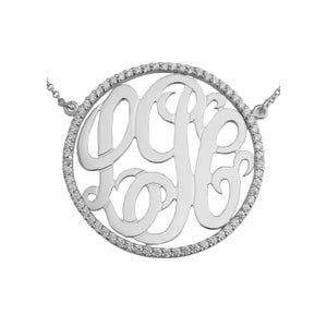 Könnte beinhalten: Eine silberne Halskette mit einem runden Anhänger, der ein Monogramm-Design zeigt, das von einem Kreis aus kleinen, klaren Steinen umgeben ist.