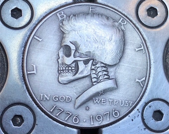 May include: A silver dollar coin with a skull profile on the front. The coin has the words "Liberty", "In God We Trust", "1776", and "1976" inscribed on it.