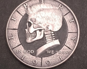 May include: Silver coin with a skull design. The skull is wearing a hat and has a skeletal neck. The coin has the words "Liberty", "In God We Rust", and "1999" inscribed on it.