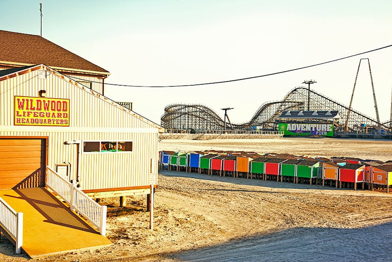 May include: A beige building with a sign that reads "Wildwood Lifeguard Headquarters". In the background, a roller coaster and a tall tower are visible. There are colourful beach huts in the foreground.