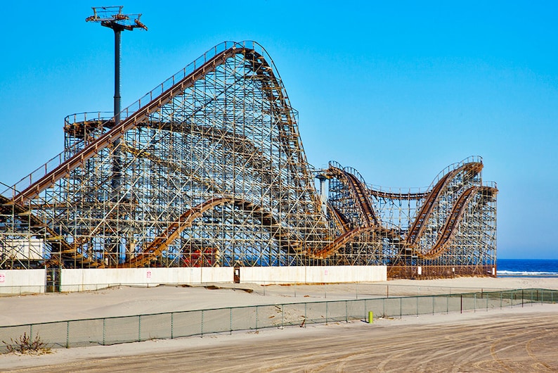 Puede incluir: Una gran monta&ntilde;a rusa de madera oxidada se alza en una playa de arena con un cielo azul y el oc&eacute;ano al fondo.