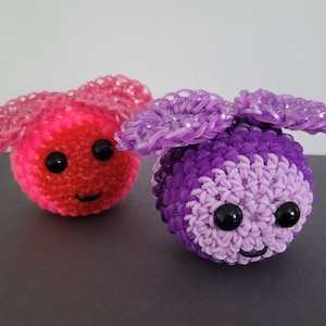 May include: Two handmade, colorful loom band creatures. One is pink and red, the other is purple and lavender. Both have black eyes and smiling faces. The toys are round with looped ears.