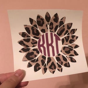 May include: A square decal featuring a sunflower design with petals in a brown and tan leopard print pattern. In the center of the flower is a circular maroon monogram containing the letters KKT in a bold, vertical, serif-style font.